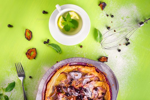 Good morning, homemade Crumble pie with black currants, plums and powdered sugar for breakfast, mint tea with lemon, fork, whisk, dried plum, mint leaves on wooden green table, top view, composition