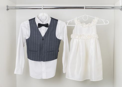 white  boy's shirt with black bow tie, vest and white elegant a little girl  dress hanging on hangers in the white wardrobe, clothes for the holiday, ceremony, wedding
