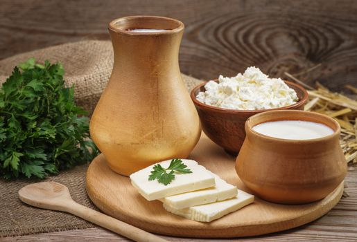 Rustic natural dairy products, whole milk in a clay jug and a cup, cottage cheese in a bowl, cut slices of cheese, parsley, burlap, straw on the old wooden background