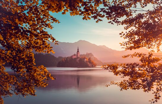 Little Island with Catholic Church in Bled Lake, Slovenia  at Sunrise with Castle and Mountains in Background. Autumn Filter. Tree Leaves Border. Natural Frame.