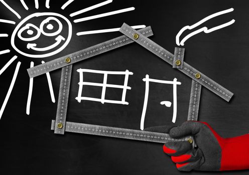 Hand holding a metal meter ruler in the shape of house with sun, door, window and smoke from the chimney. On a blackboard. Concept of house project 