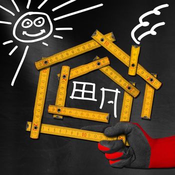 Hand holding a wooden meter ruler in the shape of house with sun, door, window and smoke from the chimney. On a blackboard. Concept of house project 