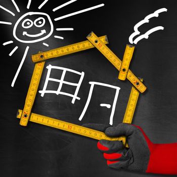 Hand holding a wooden meter ruler in the shape of house with sun, door, window and smoke from the chimney. On a blackboard. Concept of house project 