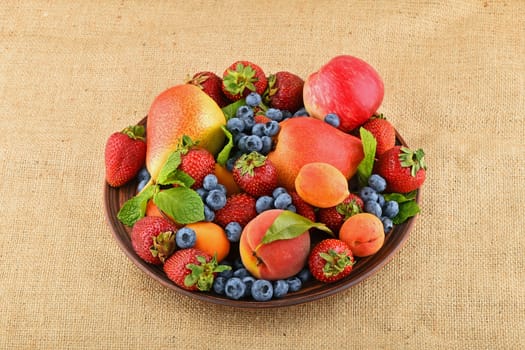 Mellow fresh summer fruits and berries mix with mint leaves in ceramic plate on burlap jute canvas, strawberries, blueberries, apricots, peach, apple and pear