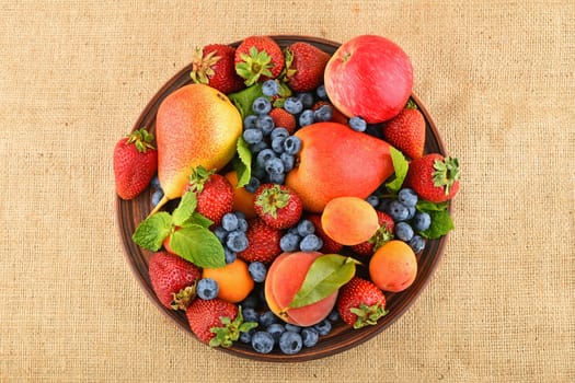 Mellow fresh summer fruits and berries mix with mint leaves in ceramic plate on burlap jute canvas, strawberries, blueberries, apricots, peach, apple and pear