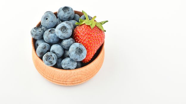 Handful of fresh blueberries and one strawberry in handmade wooden bowl isolated on white