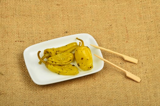Mediterranean snack of four pickled green pepper in white plate with two skewers on burlap jute canvas