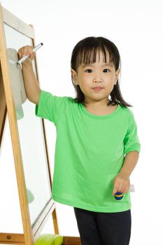 Happy cute Asian Chinese toddler girl drawing or writting with marker pen on a blank whiteboard at home, preschool, daycare or kindergarten in plain white isolated background.