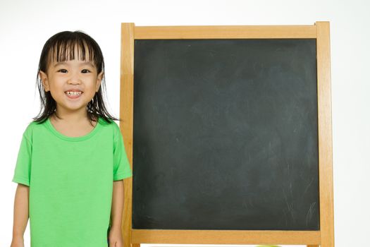 Happy cute Asian Chinese toddler girl standing beside a blank blackboard at home, preschool, daycare or kindergarten in plain white isolated background.
