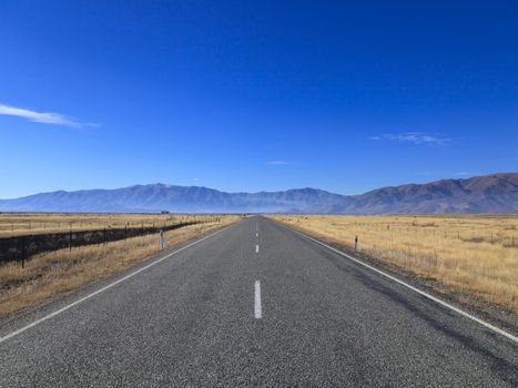 Straight empty Country Road leading to Mountains lining the horizon on sunny day, Otago region, South Island, New Zealand.