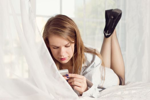 girl lies in a man's shirt on the bed with a smartphone in the hands of