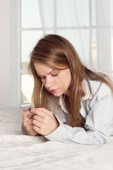 girl lies in a man's shirt on the bed with a smartphone in the hands of