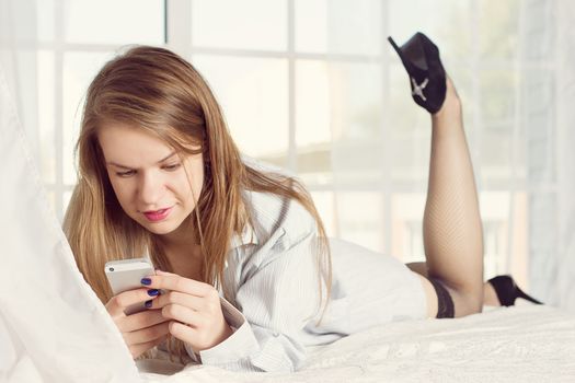 girl lies in a man's shirt on the bed with a smartphone in the hands of