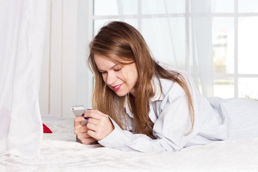 girl lies in a man's shirt on the bed with a smartphone in the hands of