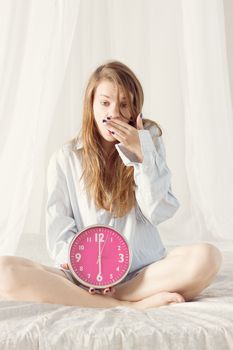 girl is sitting on the bed with big pink clock. 6 a.m