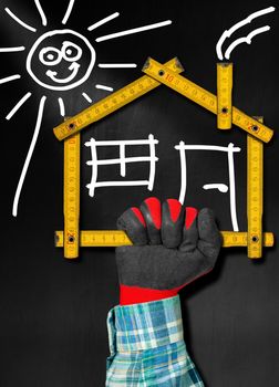 Hand holding a wooden meter ruler in the shape of house with sun, door, window and smoke from the chimney. On a blackboard. Concept of house project