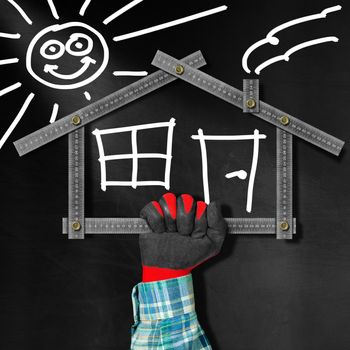 Hand holding a metal meter ruler in the shape of house with sun, door, window and smoke from the chimney. On a blackboard. Concept of house project 