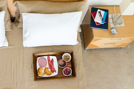 A tasty healthy breakfast in bed with coffee, fresh berries, bacon and croissants alongside a bedside cabinet with reading books for a relaxing weekend lie in bed