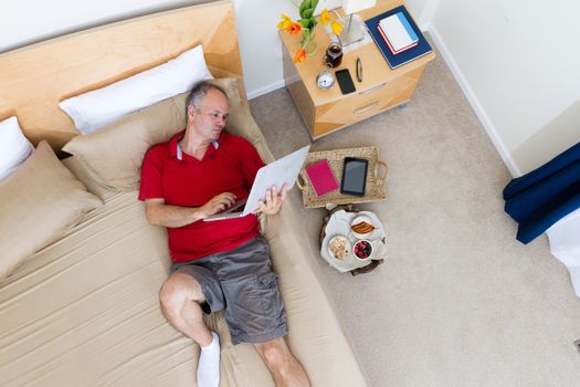 High Angle View of Mature Man Lying on Bed with Laptop in Hotel Room with Remnants of Breakfast Tray and Personal Effects on Floor