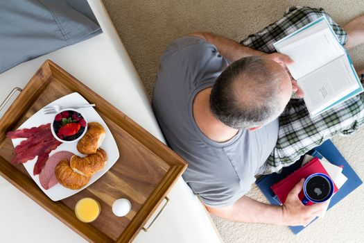 High Angle View of Mature Man in Pajamas Reading Novel and Enjoying Cup of Coffee While Sitting on Floor Leaning Against Bed with Breakfast Tray Resting on Top, a Quiet Sunday Morning