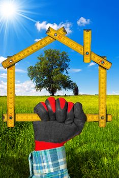 Hand with work glove holding a wooden meter ruler in the shape of house, in the countryside with green tree. Concept of ecological house project 