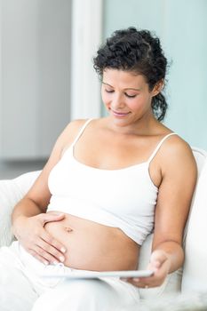 Happy pregnant woman sitting with tablet pc on sofa at home