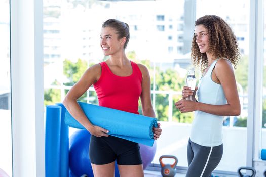 Cheerful women in fitness studio holding exercise mat and water bottle