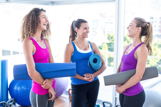 Women holding exercise mat and smiling while looking at each other in fitness studio