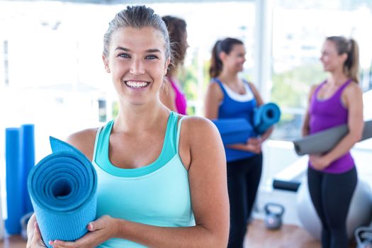 Portrait of cheerful woman holding exercise mat with friends at fitness studio