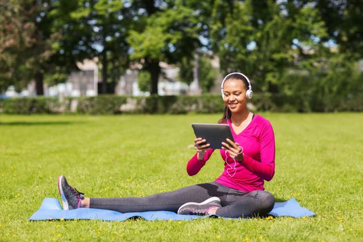 fitness, park, technology and sport concept - smiling african american woman with tablet pc computer and headphones on mat outdoors