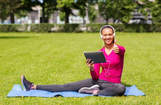 fitness, park, technology and sport concept - smiling african american woman with tablet pc and headphones showing thumbs up outdoors