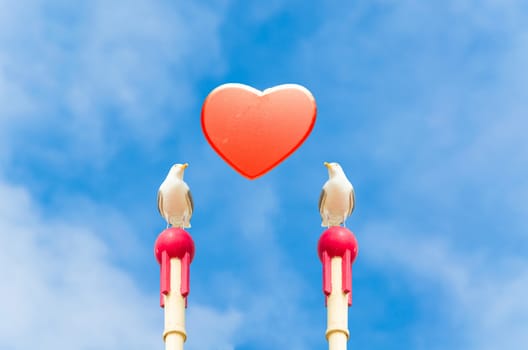 Two large seagull sitting on a wooden post. In the middle of a red heart in front of blue sky. Symbol of love.
