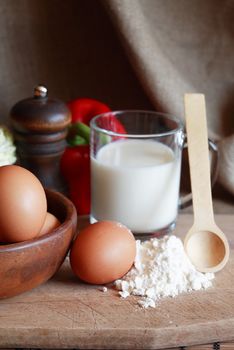 Bowl with raw eggs and meal near cheese and milk on wooden cutting board