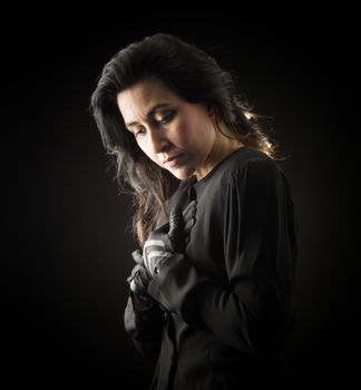Brunette woman in black standing in front of black backdrop, clasping her hands to her chest and looking down with a serious, sad expression on her face