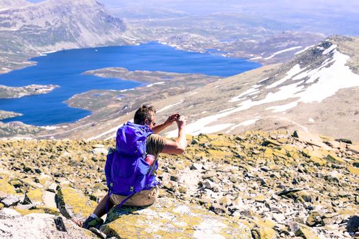 Hiker with big traveling rucksack taking pictures on the phone on the mountain trail, adventure travel and discovery