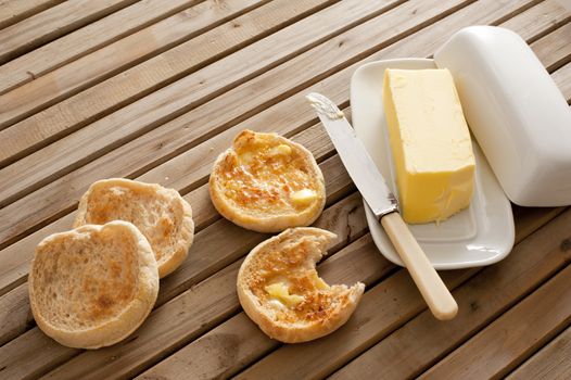 Fresh toasted hot English muffins, one with a bite missing, on a wooden slatted table with a pat of butter in a butter dish