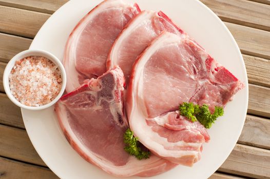 Overhead view of four raw pork cutlets with their rind arranged on a plate ready for cooking with a small dish of natural rock or sea salt on a wooden table