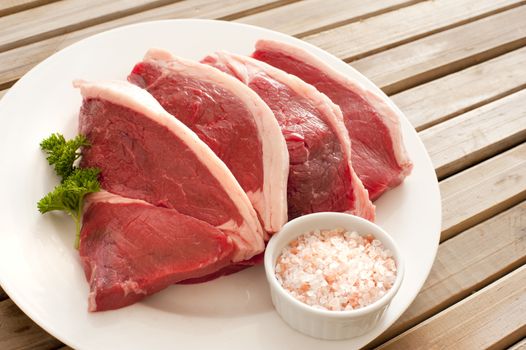 Four uncooked succulent beef steaks arranged ready for cooking on a plate with a bowl of rock salt and parsley garnish