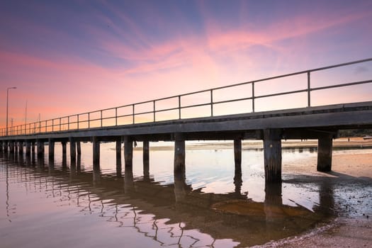 A long wooden jetty with the tide out at sunset. Boats just visible in the background.