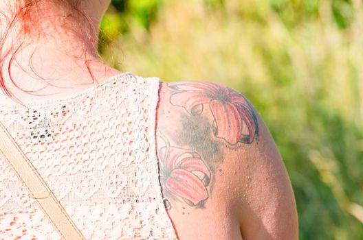 Rotharrige young woman with flowers tattoo on her right shoulder blade. In the background, meadow and trees.