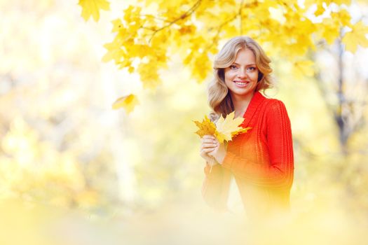 Cheerful young woman sitting on the ground covered with dry autumnal foliage in beautiful park