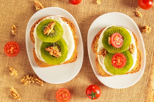 Toast with kiwi, cheese, walnuts and cherry tomato  on a piece of sackcloth with walnuts and cherry tomatoes around. View from above