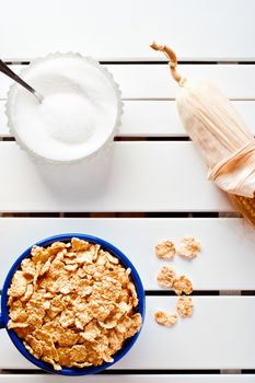 Cornflakes in a blue bowl on white wooden table with cob and sugar