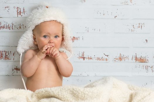 Naked baby girl sitting in a warm interior wearing white cap. Rustic wall in the back.
