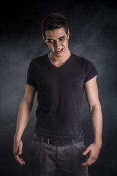 Portrait of a Young Vampire Man with Black T-Shirt, Looking at the Camera, on a Dark Smoky Background.