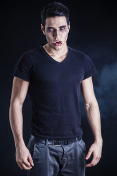 Portrait of a Young Vampire Man with Black T-Shirt, Looking at the Camera, on a Dark Smoky Background.