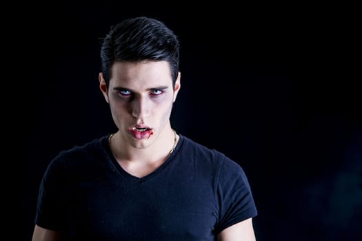 Portrait of a Young Vampire Man with Black T-Shirt, Looking at the Camera, on a Dark Smoky Background.