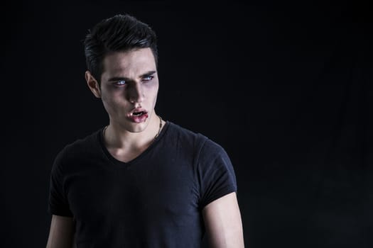 Portrait of a Young Vampire Man with Black T-Shirt, Looking at the Camera, on a Dark Smoky Background.