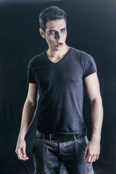 Portrait of a Young Vampire Man with Black T-Shirt, Looking at the Camera, on a Dark Smoky Background.