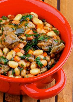 Tasty White Beans Stew with Boiled Beef and Greens in Rustic Red Pot closeup on Wooden Plank background